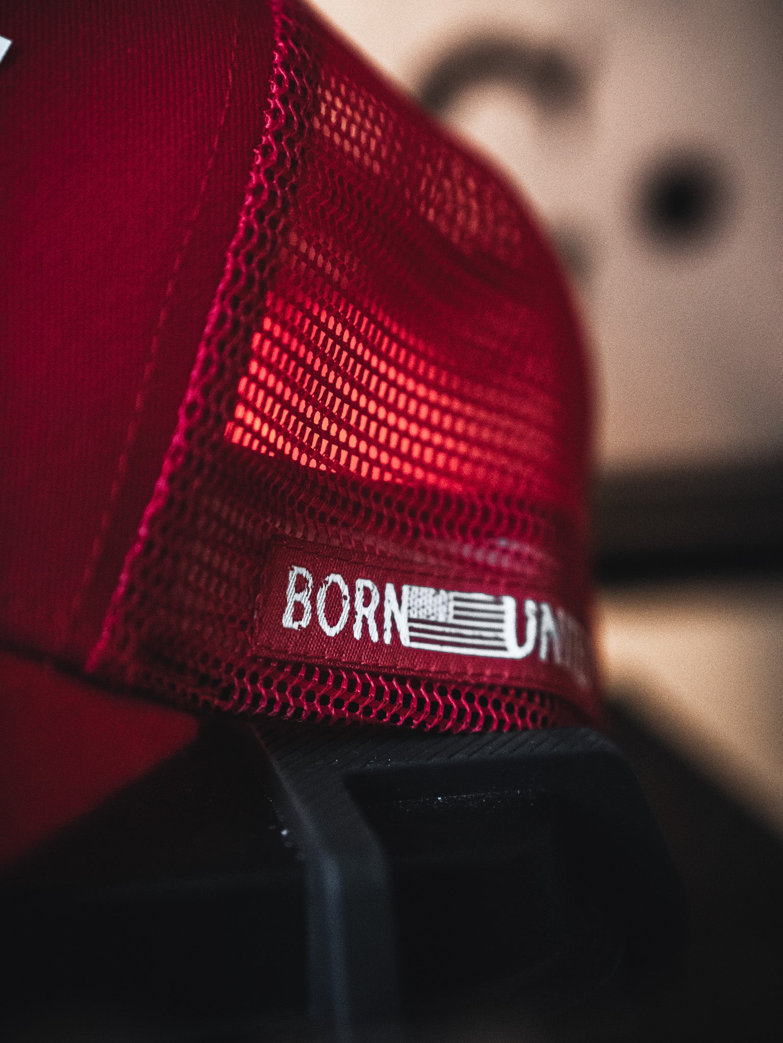 A close-up of the Live Like Charlie Trucker Snapback by Born United Private Label, featuring red mesh, a 5-panel curved brim, and "BORN" with part of a "U" printed on the mesh. An abstract shape appears blurred in the background.