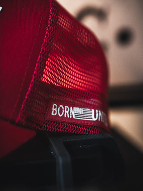 A close-up of the Live Like Charlie Trucker Snapback by Born United Private Label, featuring red mesh, a 5-panel curved brim, and "BORN" with part of a "U" printed on the mesh. An abstract shape appears blurred in the background.
