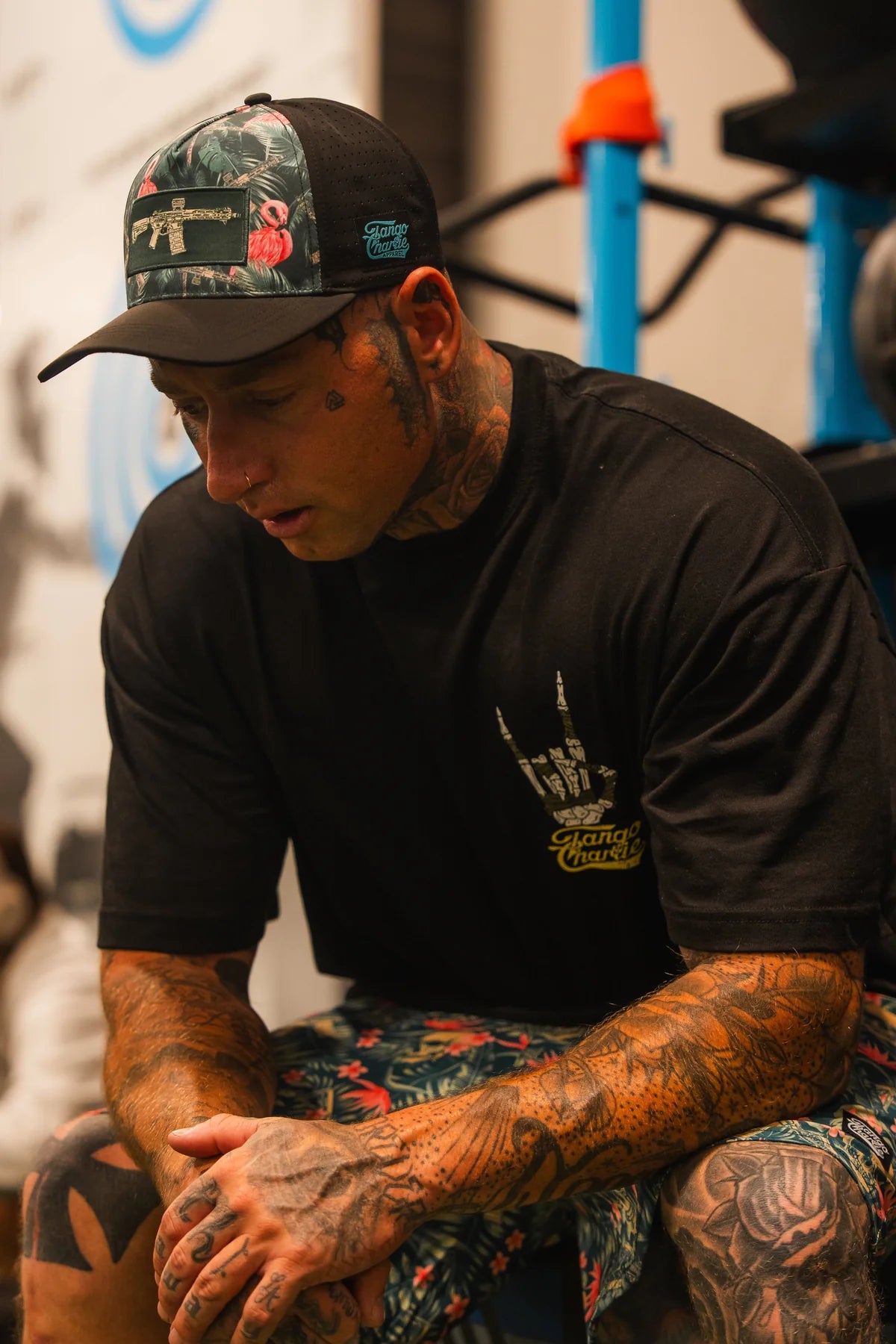 A heavily tattooed man wearing the Tango Charlie Apparel Gain Gang Premium Oversized Tee sits with his hands clasped, looking down thoughtfully.