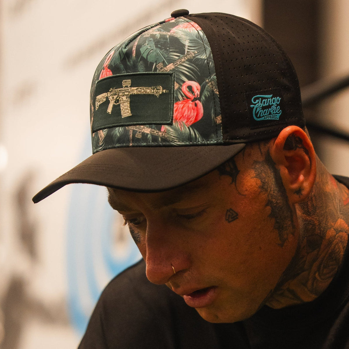 A person with face tattoos wears the Loaded Flamingo Hydro Snapback Hat by Tango Charlie Apparel, which has a black and floral design with a gold gun patch. They are looking down, with a blurred background behind them.