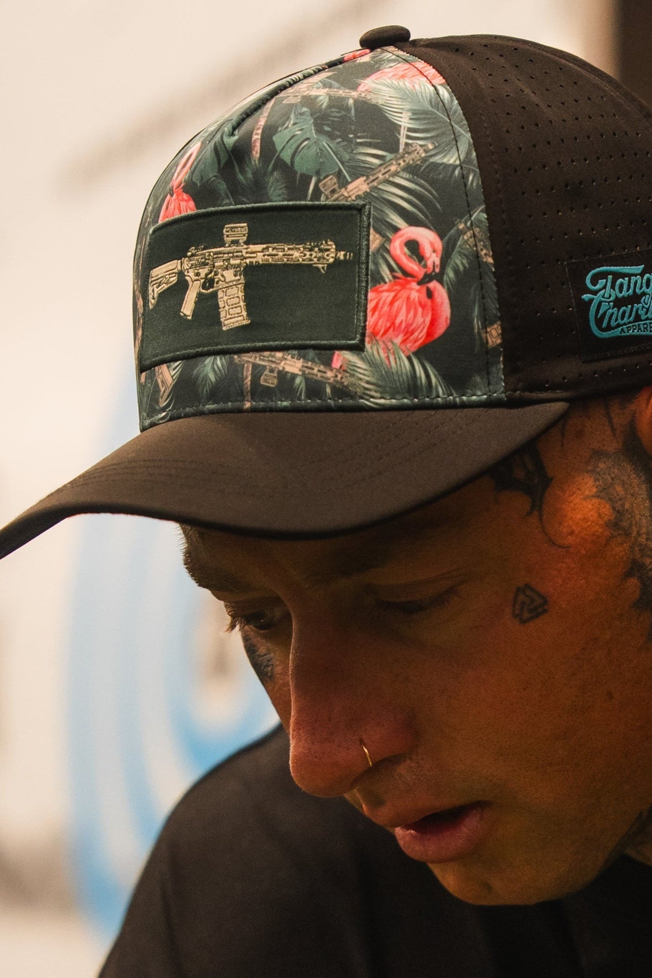 A person with face tattoos wears the Loaded Flamingo Hydro Snapback Hat by Tango Charlie Apparel, which has a black and floral design with a gold gun patch. They are looking down, with a blurred background behind them.