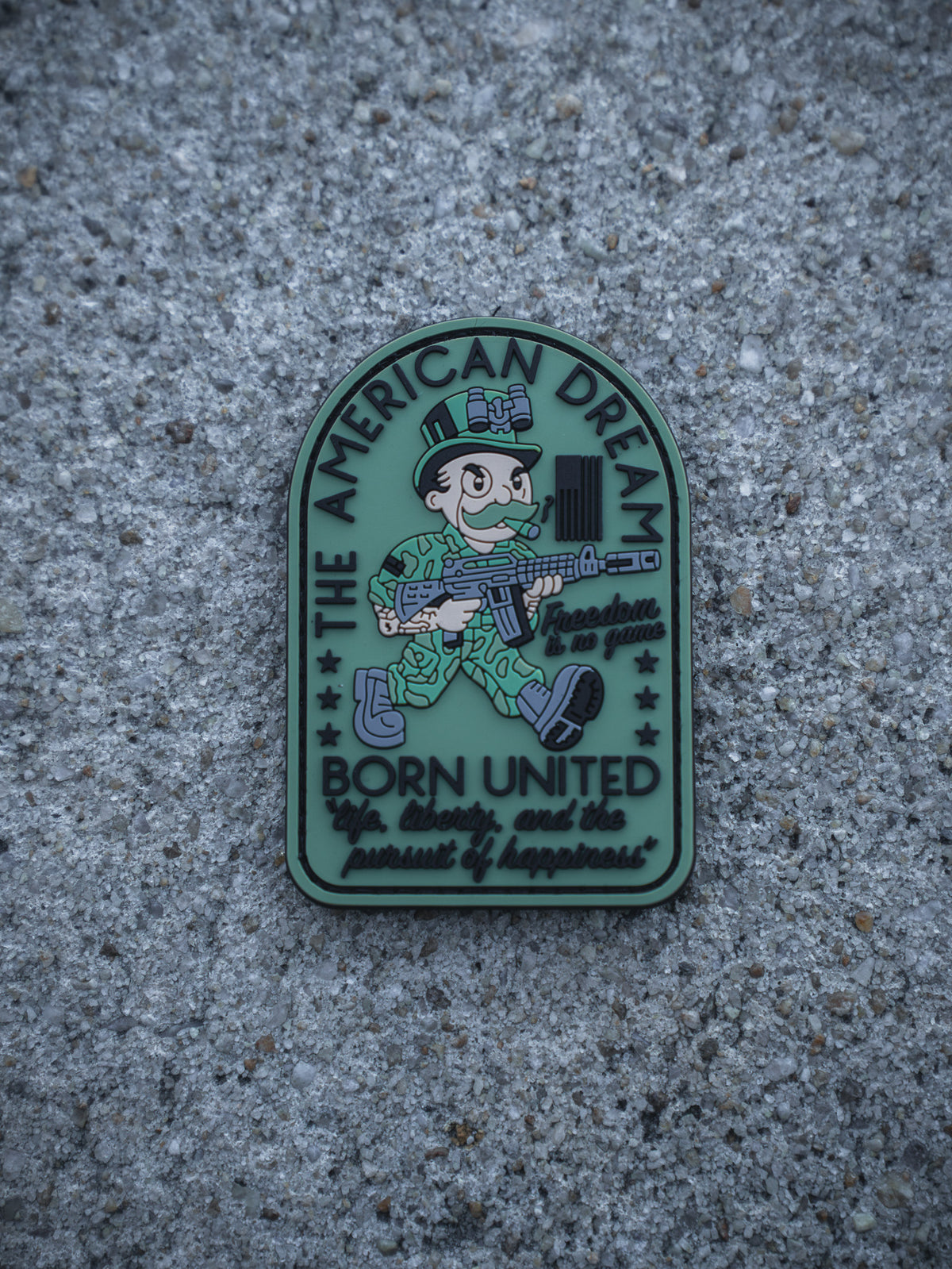 The American Dream PVC Patch by Born United Private Label displays a cartoon soldier with a rifle on a gray background, with "The American Dream, Born United, life, liberty, and the pursuit of happiness" plus stars and a flag.