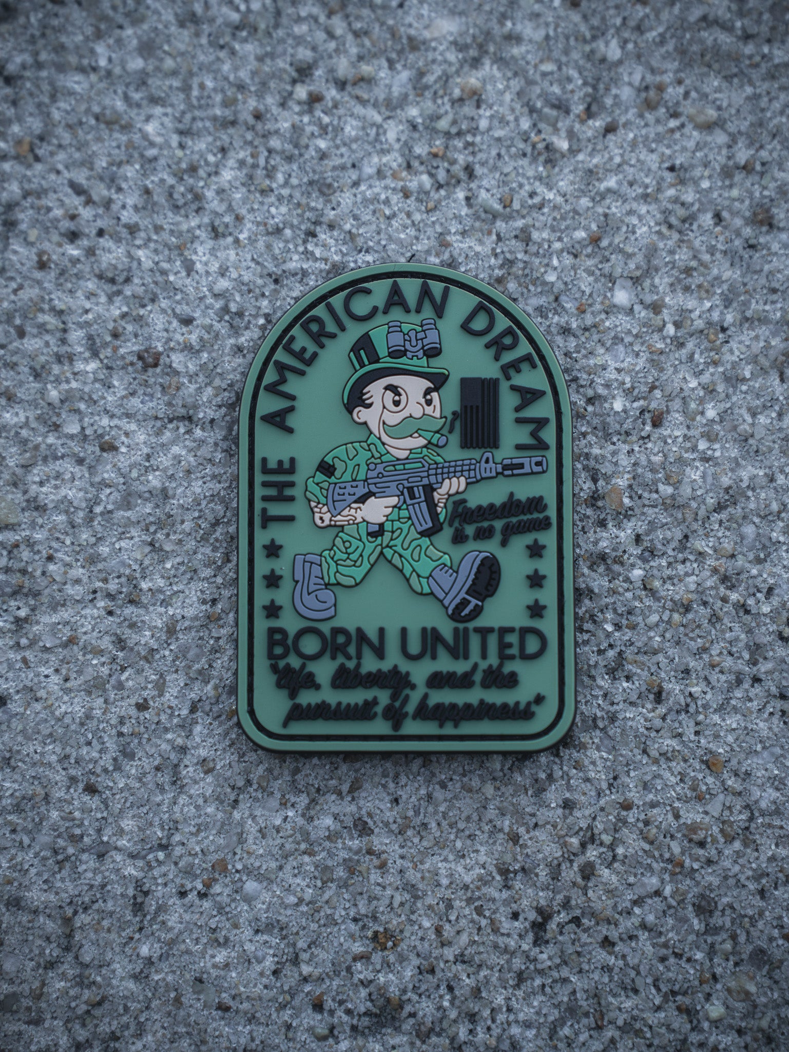 The American Dream PVC Patch by Born United Private Label displays a cartoon soldier with a rifle on a gray background, with "The American Dream, Born United, life, liberty, and the pursuit of happiness" plus stars and a flag.