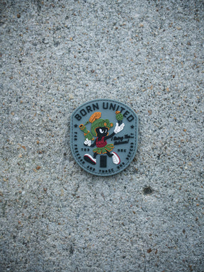 The Kaboom! PVC Patch by Born United Private Label features a colorful cartoon character in Roman armor with sneakers, sword, and helmet, set on a speckled concrete background. “BORN UNITED” is displayed at the top.