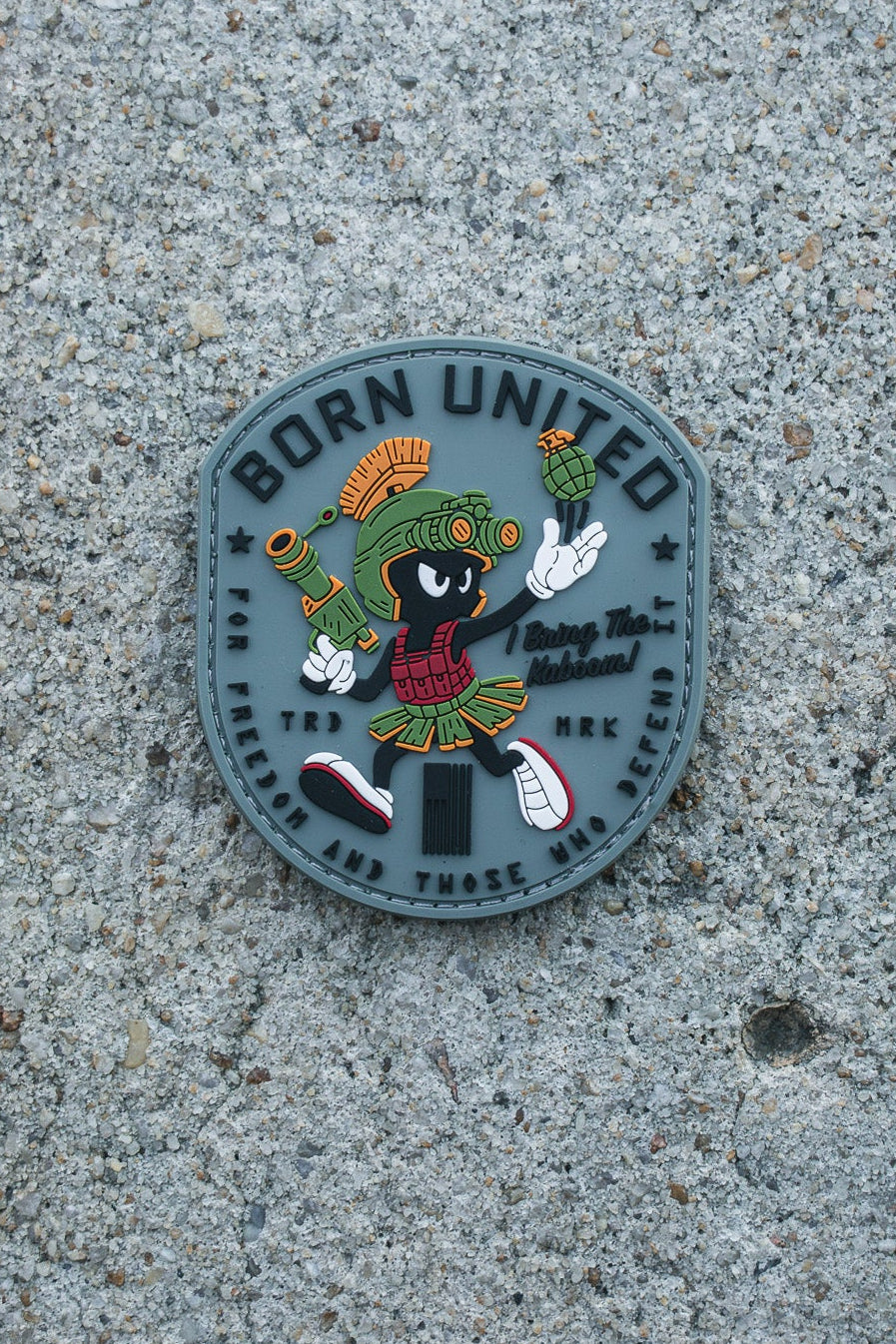 The Kaboom! PVC Patch by Born United Private Label features a colorful cartoon character in Roman armor with sneakers, sword, and helmet, set on a speckled concrete background. “BORN UNITED” is displayed at the top.
