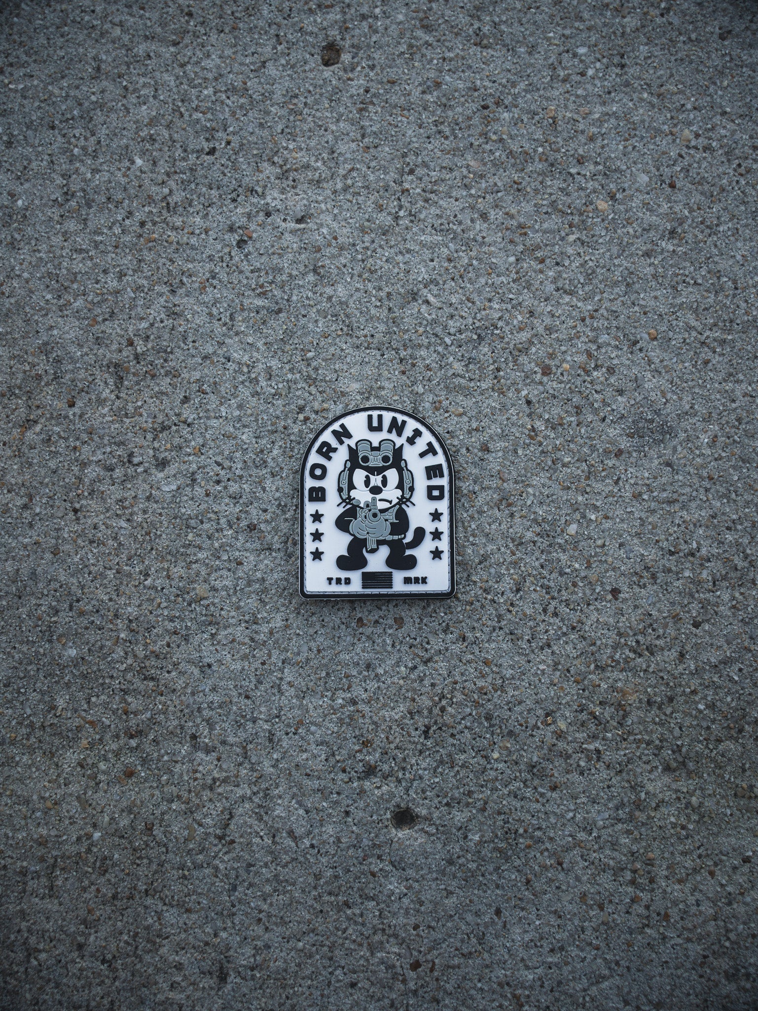 A black and white Foxtrot Felix PVC Patch by Born United Private Label sits on gray concrete, showing a cartoon cat hugging a bear with "BORN UNITED" and stars above, and "TKD BRK" below the characters.