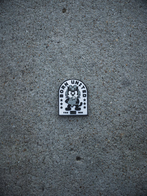 A black and white Foxtrot Felix PVC Patch by Born United Private Label sits on gray concrete, showing a cartoon cat hugging a bear with "BORN UNITED" and stars above, and "TKD BRK" below the characters.