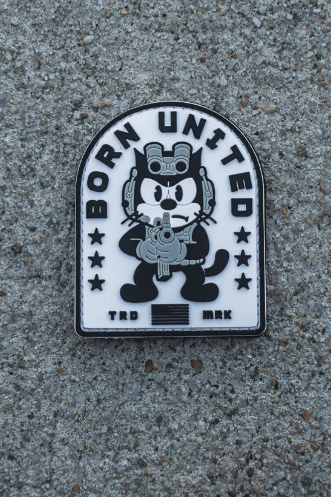 A black and white Foxtrot Felix PVC Patch by Born United Private Label sits on gray concrete, showing a cartoon cat hugging a bear with "BORN UNITED" and stars above, and "TKD BRK" below the characters.