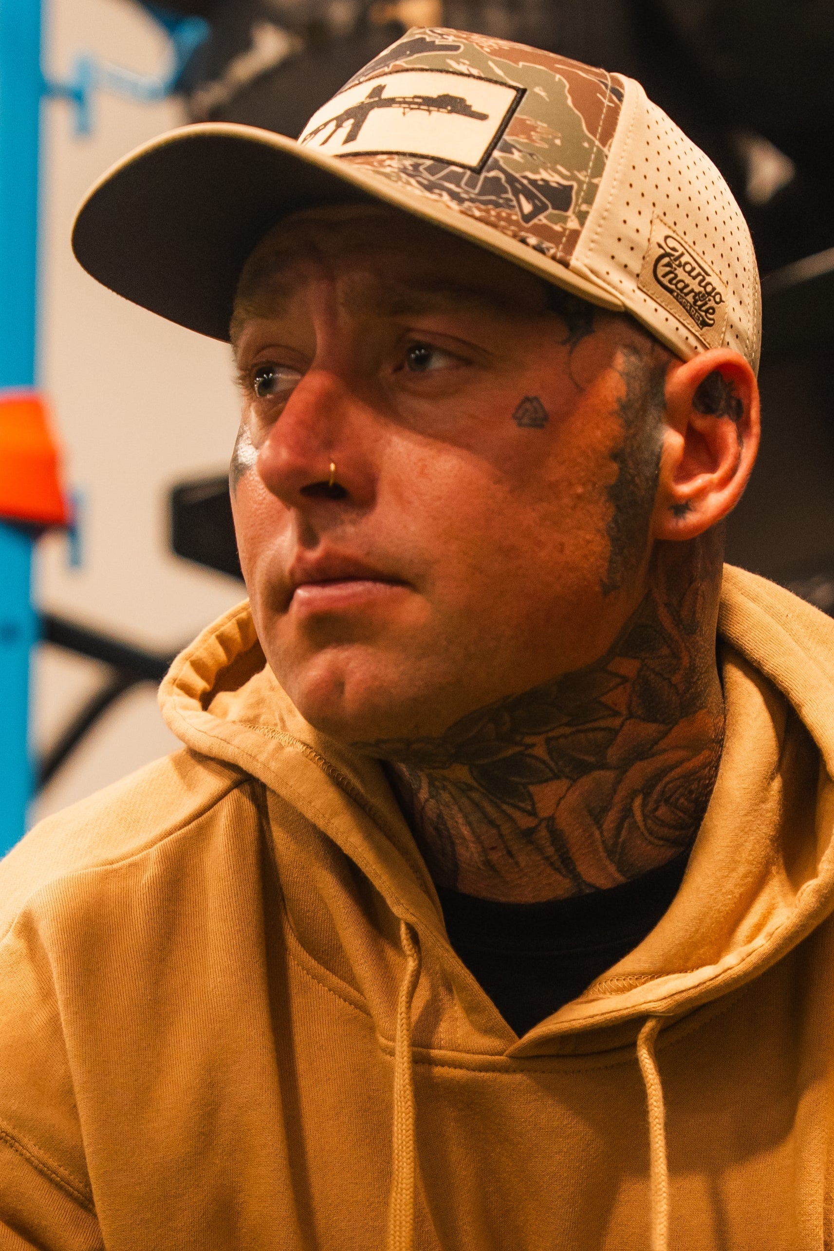 A man wearing a tan hoodie and the Tango Charlie Apparel "Snipes & Stripes Hydro" snapback hat looks to the side. He has face and neck tattoos, a nose ring, and a small heart tattoo on his cheek. The background is softly blurred.