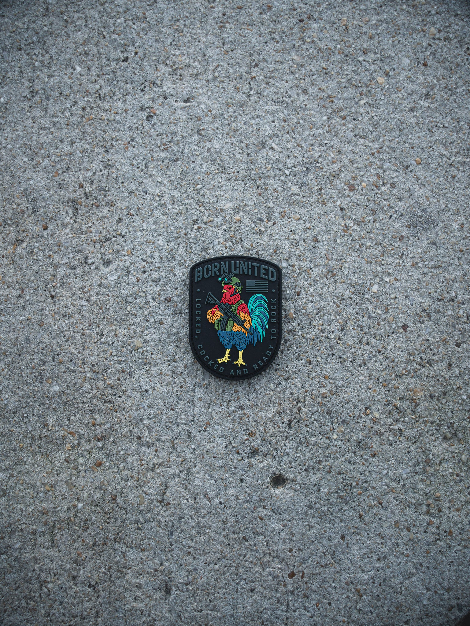 The Battle Bird PVC Patch by Born United Private Label features a colorful rooster and the words "BORN UNITED," "SOCCER," and "PHILADELPHIA" on a gray, textured concrete surface.
