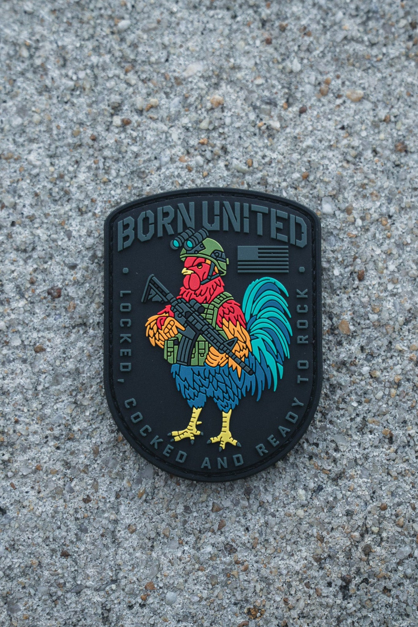 The Battle Bird PVC Patch by Born United Private Label shows a vibrant rooster in a helmet with a rifle. It reads "BORN UNITED" on top and "LOCKED, COCKED AND READY TO ROCK" below, all set against a rough concrete backdrop.