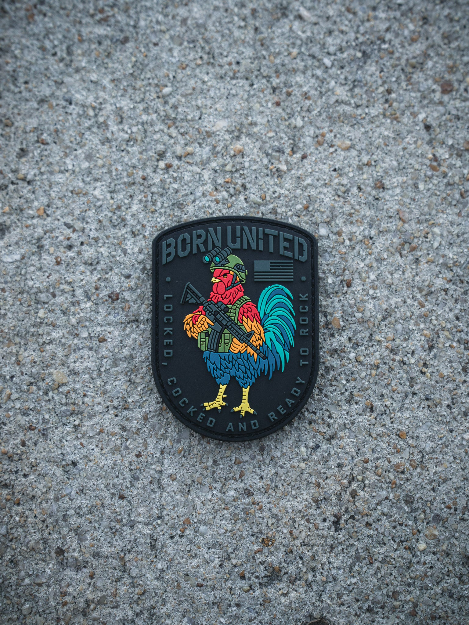 The Battle Bird PVC Patch by Born United Private Label shows a vibrant rooster in a helmet with a rifle. It reads "BORN UNITED" on top and "LOCKED, COCKED AND READY TO ROCK" below, all set against a rough concrete backdrop.