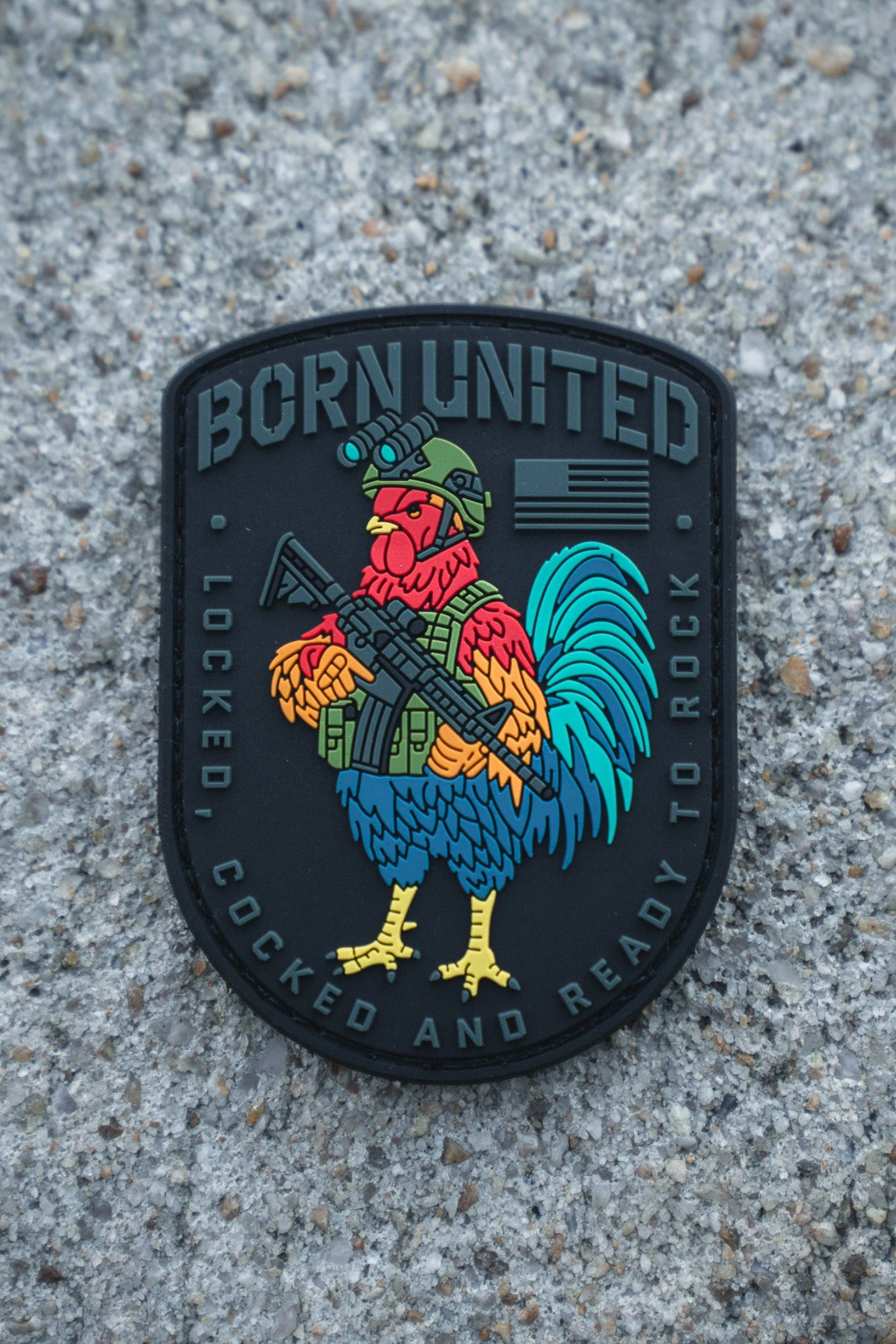 The Battle Bird PVC Patch by Born United Private Label features a colorful rooster in military gear with a rifle, “BORN UNITED” at the top, and “LOCKED, COCKED AND READY TO ROCK” at the bottom, set against a rough concrete surface.