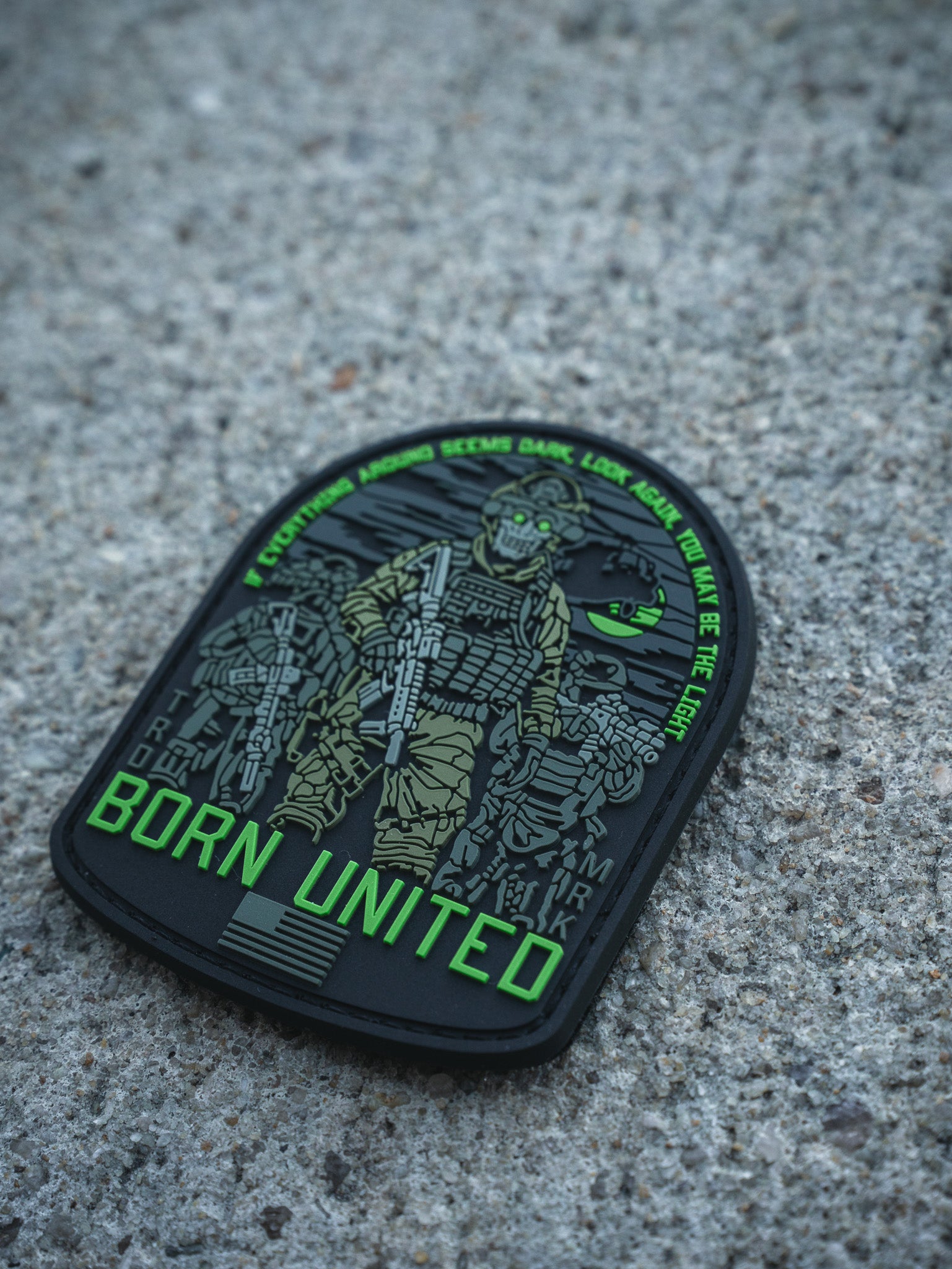 The Light in the Darkness PVC Patch by Born United Private Label features an armed soldier in night vision gear on concrete, "Born United" in bold green, and the quote: “Even if the world seems dark, look for the light. We are the light.”.