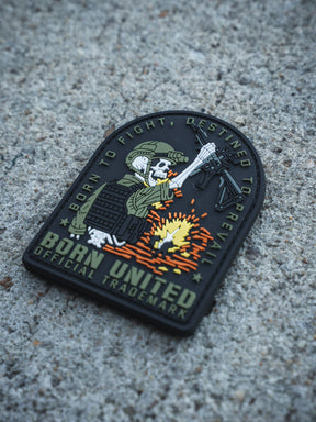 The Destined to Prevail PVC Patch by Born United Private Label shows a skeleton in combat gear with a rifle and explosion, reading “BORN TO FIGHT, DESTINED TO PREVAIL. BORN UNITED OFFICIAL TRADEMARK.” on rough concrete.