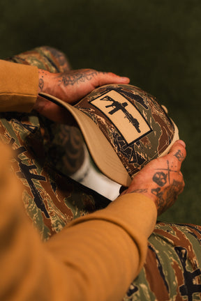 A person with tattooed hands holds the Tango Charlie Apparel Snipes & Stripes Hydro snapback hat, handmade in a camouflage pattern with a black rifle patch. They wear matching camo pants and a brown long-sleeve shirt.