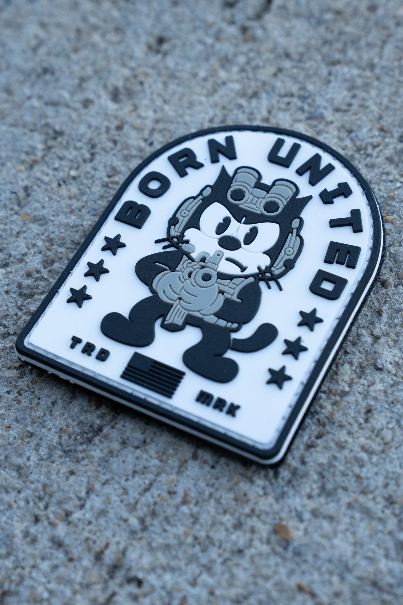 The Foxtrot Felix PVC Patch by Born United Private Label shows a cartoon cat in military gear holding a gun, set on black and white concrete with stars and "Born United" at the top, plus "TRD" and "MRK" at the bottom.