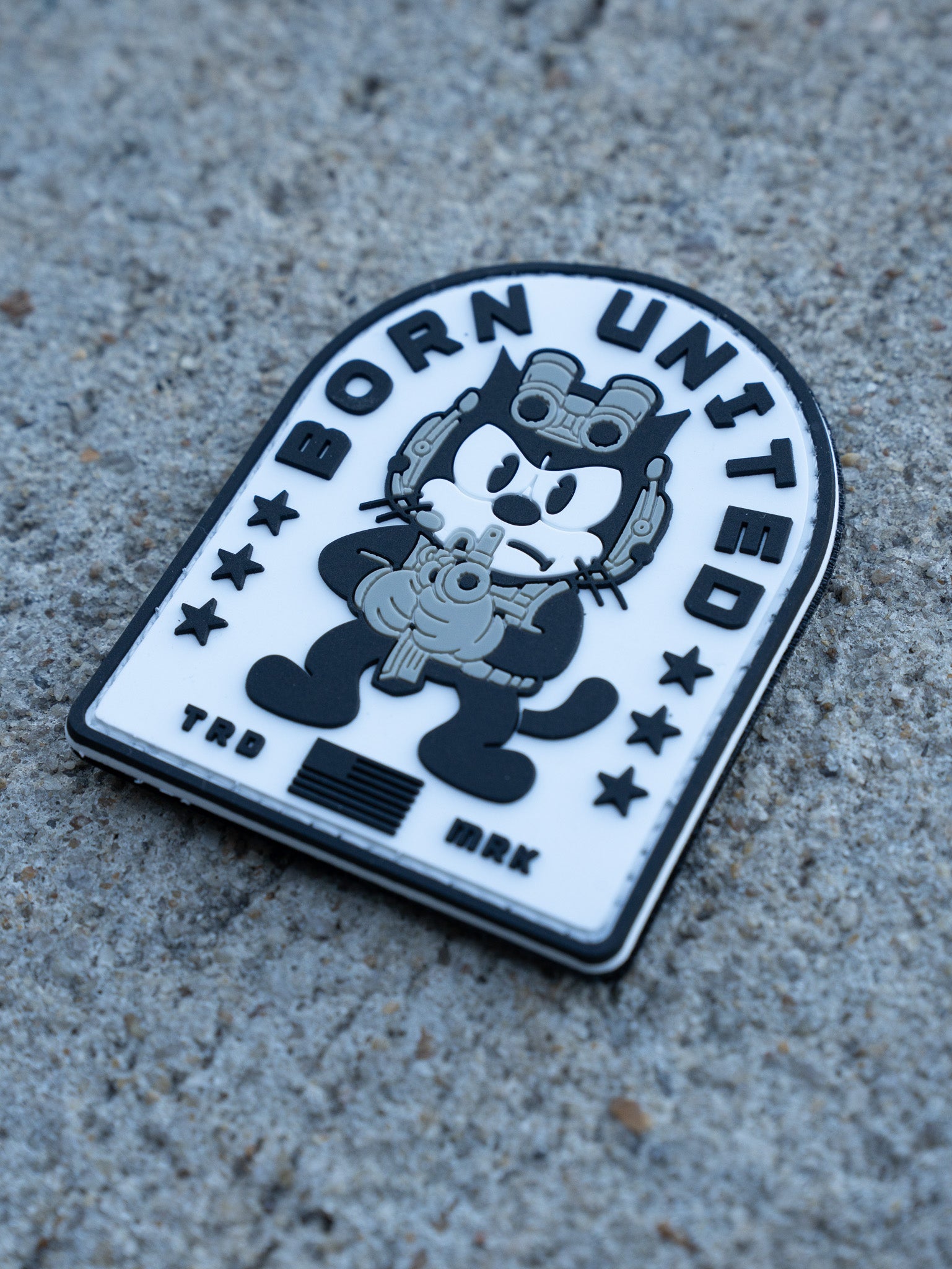 The Foxtrot Felix PVC Patch by Born United Private Label shows a cartoon cat in military gear holding a gun, set on black and white concrete with stars and "Born United" at the top, plus "TRD" and "MRK" at the bottom.