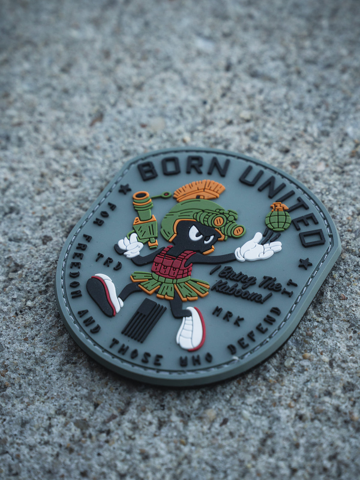 The Kaboom! PVC Patch by Born United Private Label features a cartoon alien with a blaster and grenade, wearing a Roman helmet and skirt, plus the texts "BORN UNITED", "Enjoy The Adventure", and a motto about freedom and defense on a textured surface.