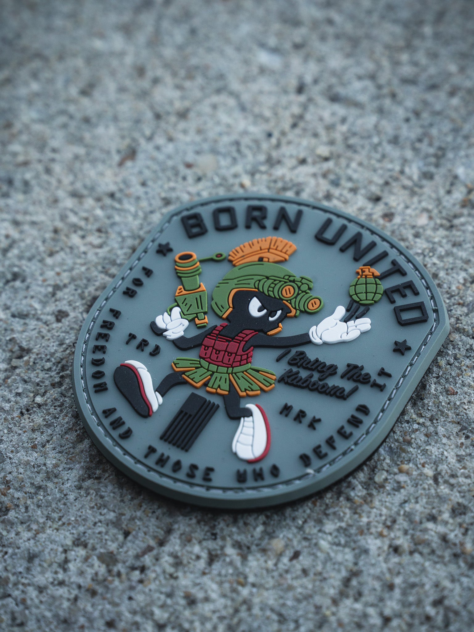 The Kaboom! PVC Patch by Born United Private Label features a cartoon alien with a blaster and grenade, wearing a Roman helmet and skirt, plus the texts "BORN UNITED", "Enjoy The Adventure", and a motto about freedom and defense on a textured surface.