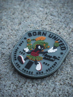 The Kaboom! PVC Patch by Born United Private Label features a cartoon alien with a blaster and grenade, wearing a Roman helmet and skirt, plus the texts "BORN UNITED", "Enjoy The Adventure", and a motto about freedom and defense on a textured surface.