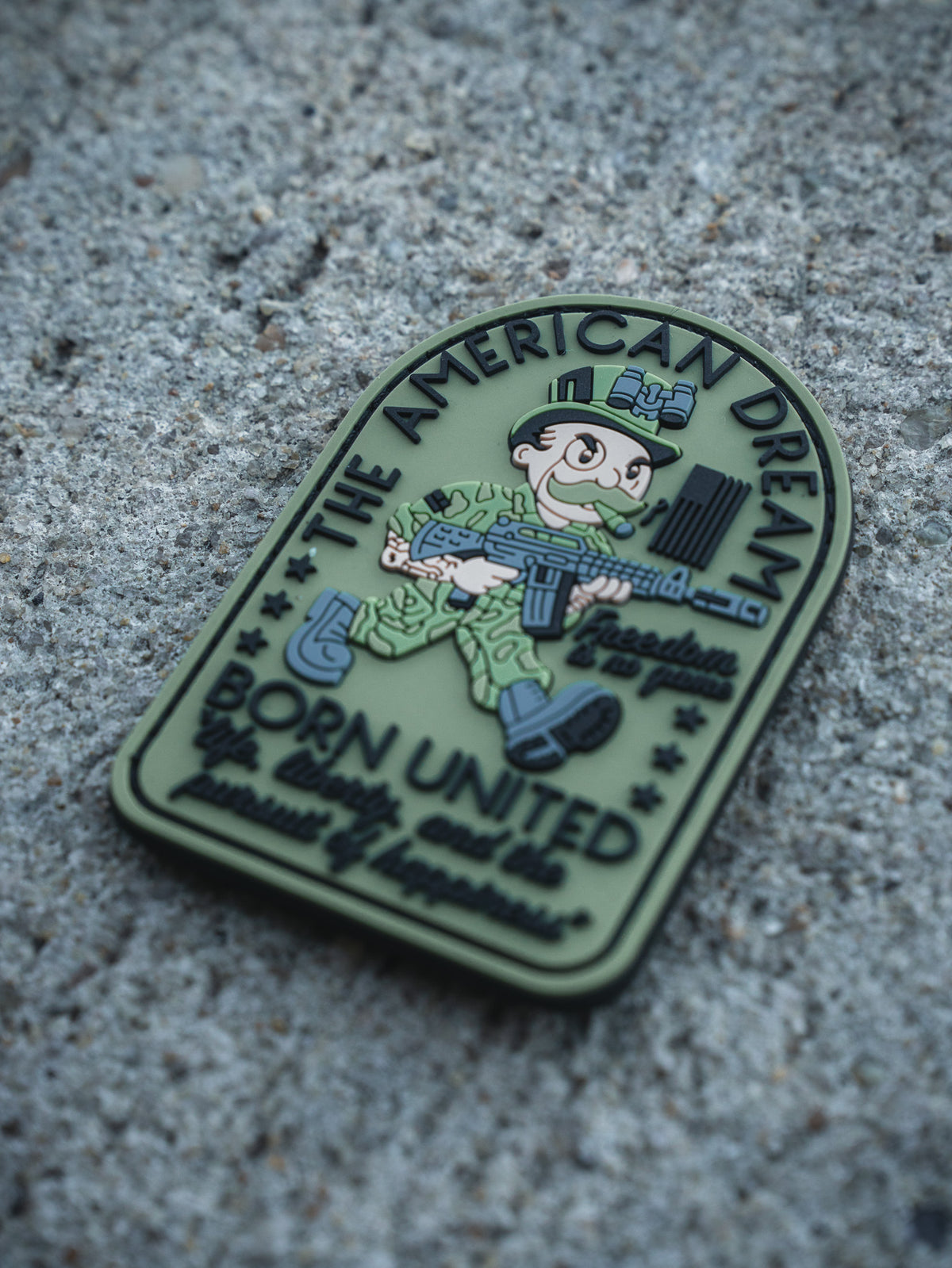 The American Dream PVC Patch by Born United Private Label features a cartoon soldier with a rifle on rough concrete, reading "THE AMERICAN DREAM," "Freedom Isn’t Free," and "BORN UNITED by unity we are powerfully unbreakable.