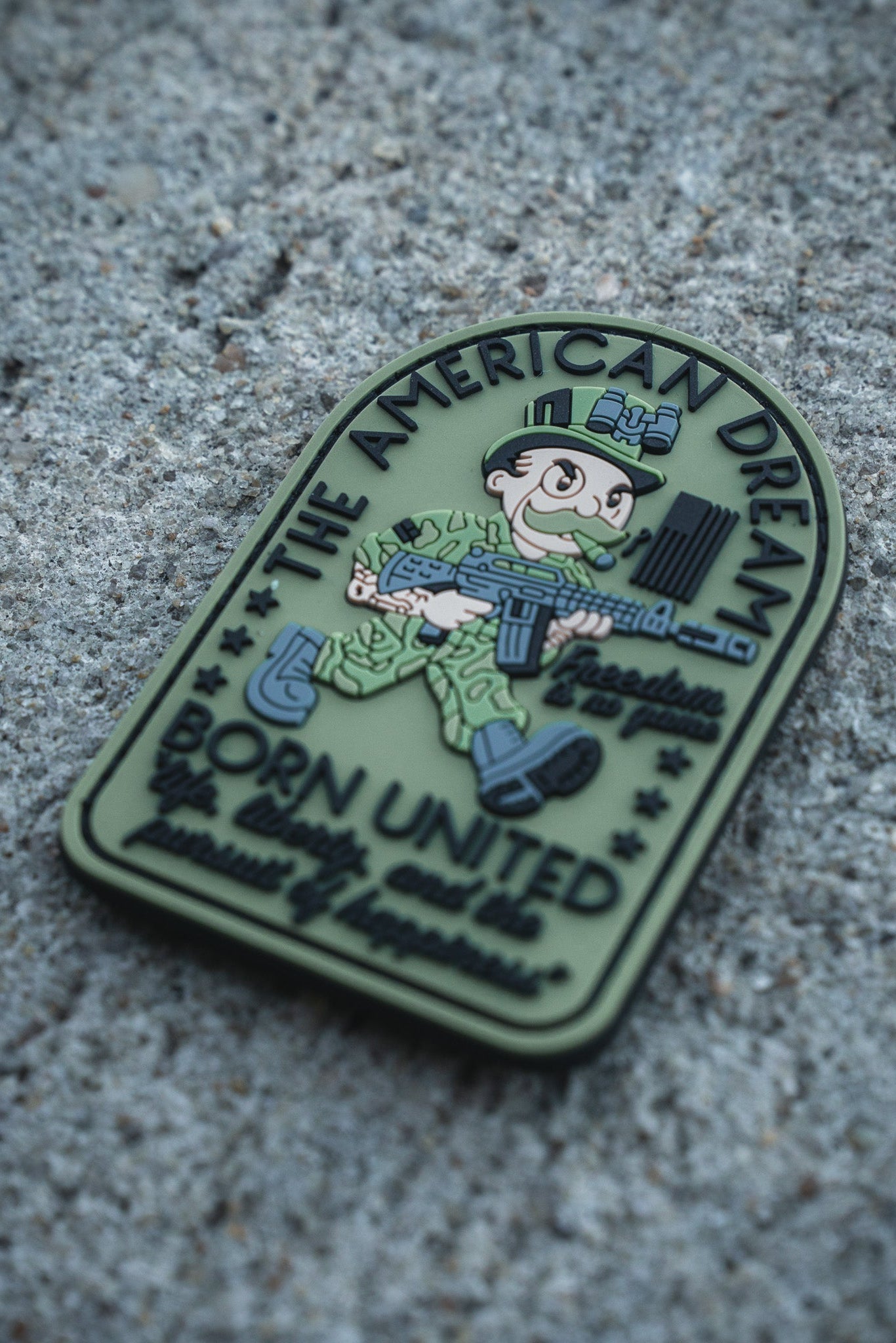 The American Dream PVC Patch by Born United Private Label features a cartoon soldier with rifle and flag, displaying “The American Dream” and “Born United” along with other patriotic phrases—perfect for those who honor service and country.