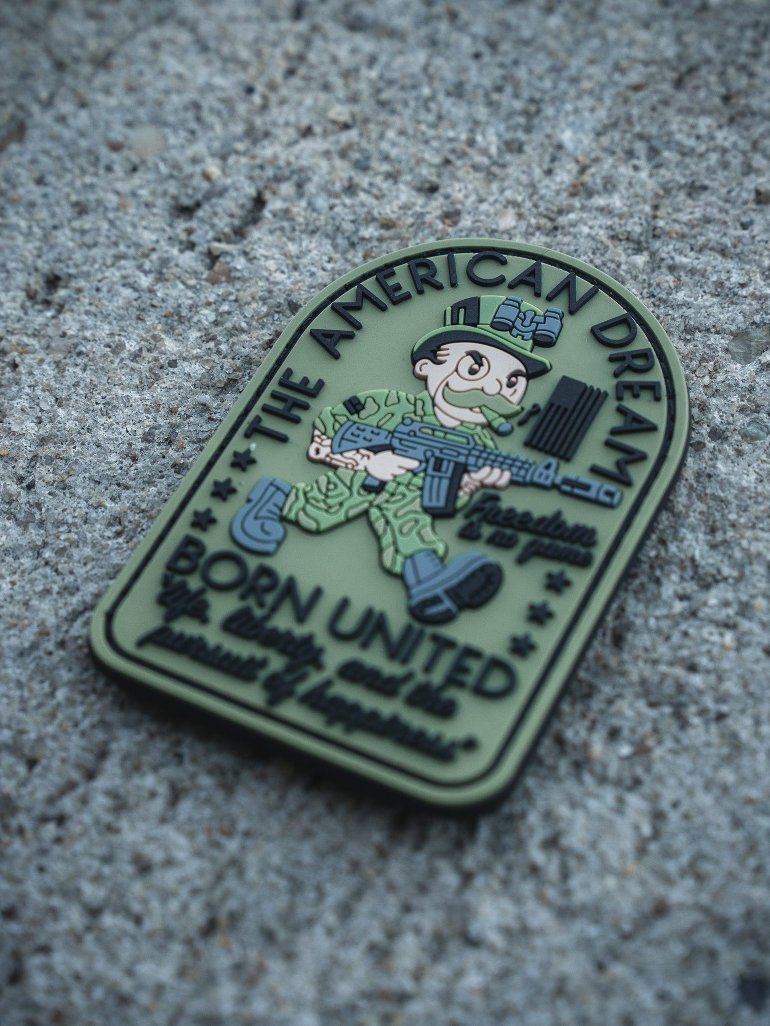 The American Dream PVC Patch by Born United Private Label features a cartoon soldier with a rifle on rough concrete, reading "THE AMERICAN DREAM," "Freedom Isn’t Free," and "BORN UNITED by unity we are powerfully unbreakable.