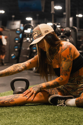 A woman with colorful tattoos wears a Tango Charlie Apparel Snipes & Stripes Hydro Snapback Hat, crop top, and patterned shorts while stretching on gym turf. A kettlebell and other equipment are visible behind her.