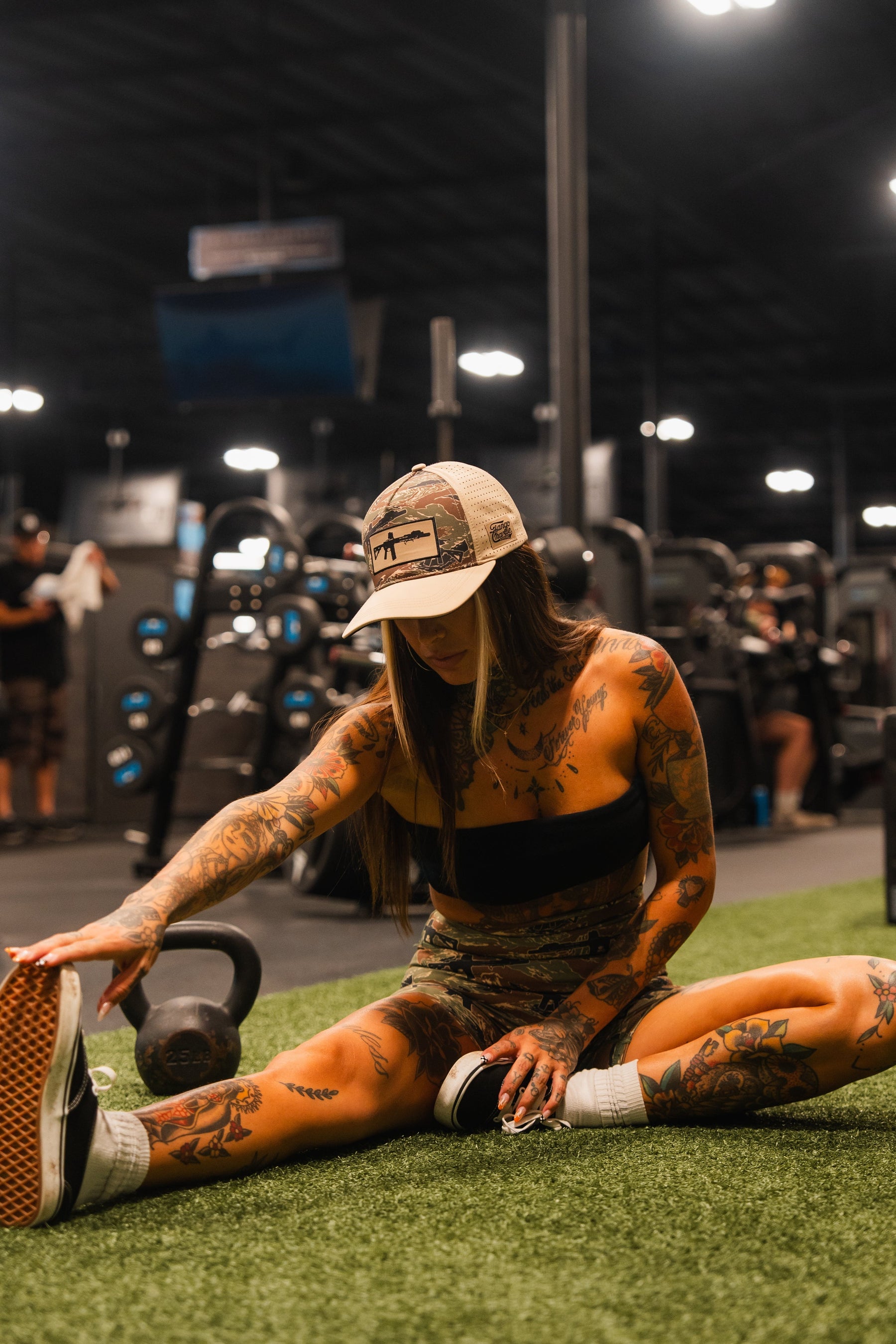 A tattooed woman stretches her leg on astroturf in a gym, wearing the Snipes & Stripes Hydro Snapback Hat by Tango Charlie Apparel, athletic clothing, and sneakers. Gym equipment and other people are visible in the background.