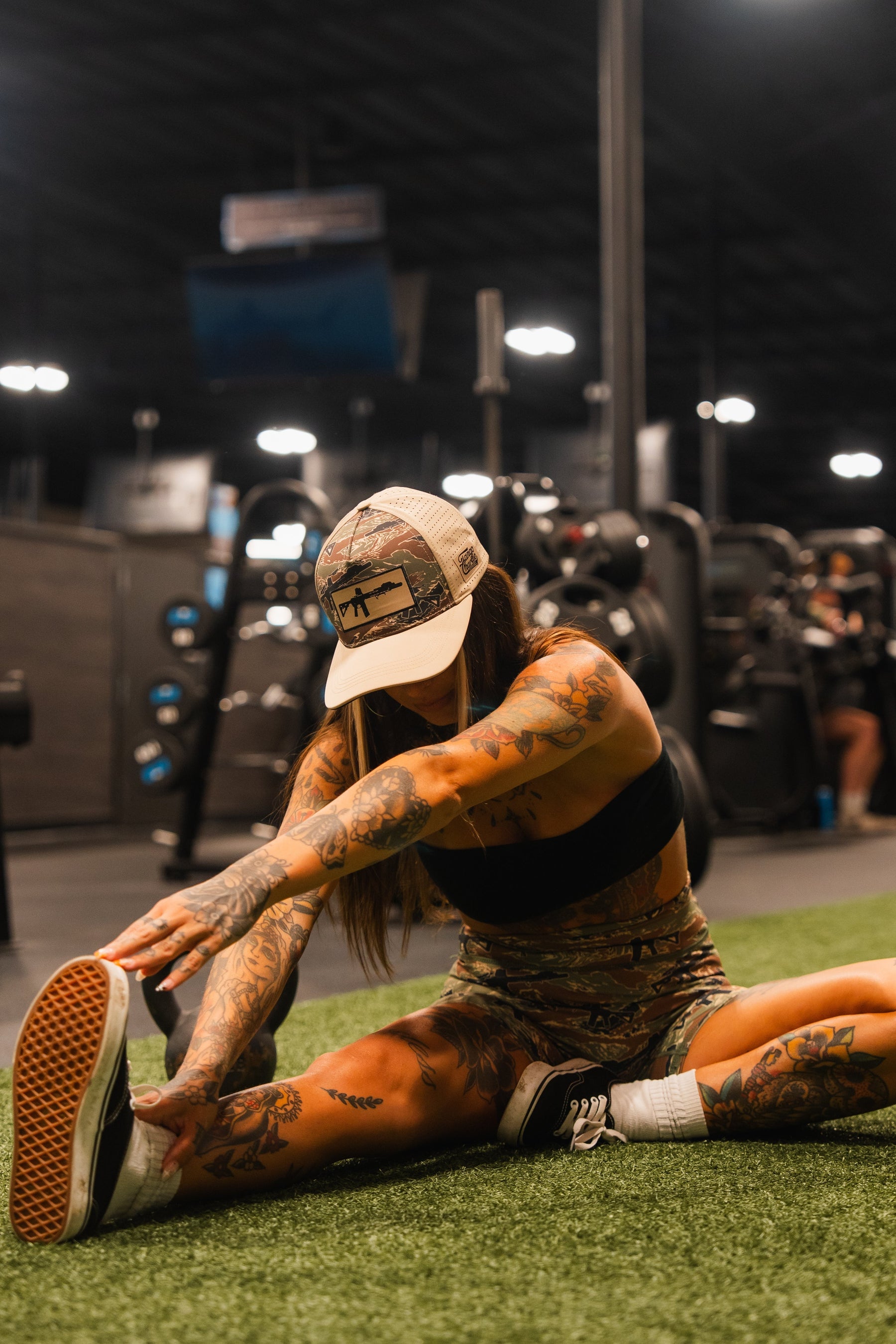 A tattooed woman wearing a Tango Charlie Apparel Snipes & Stripes Hydro Snapback Hat, shorts, and a sports bra stretches on gym turf, reaching for her foot with one leg extended, with gym equipment visible in the background.