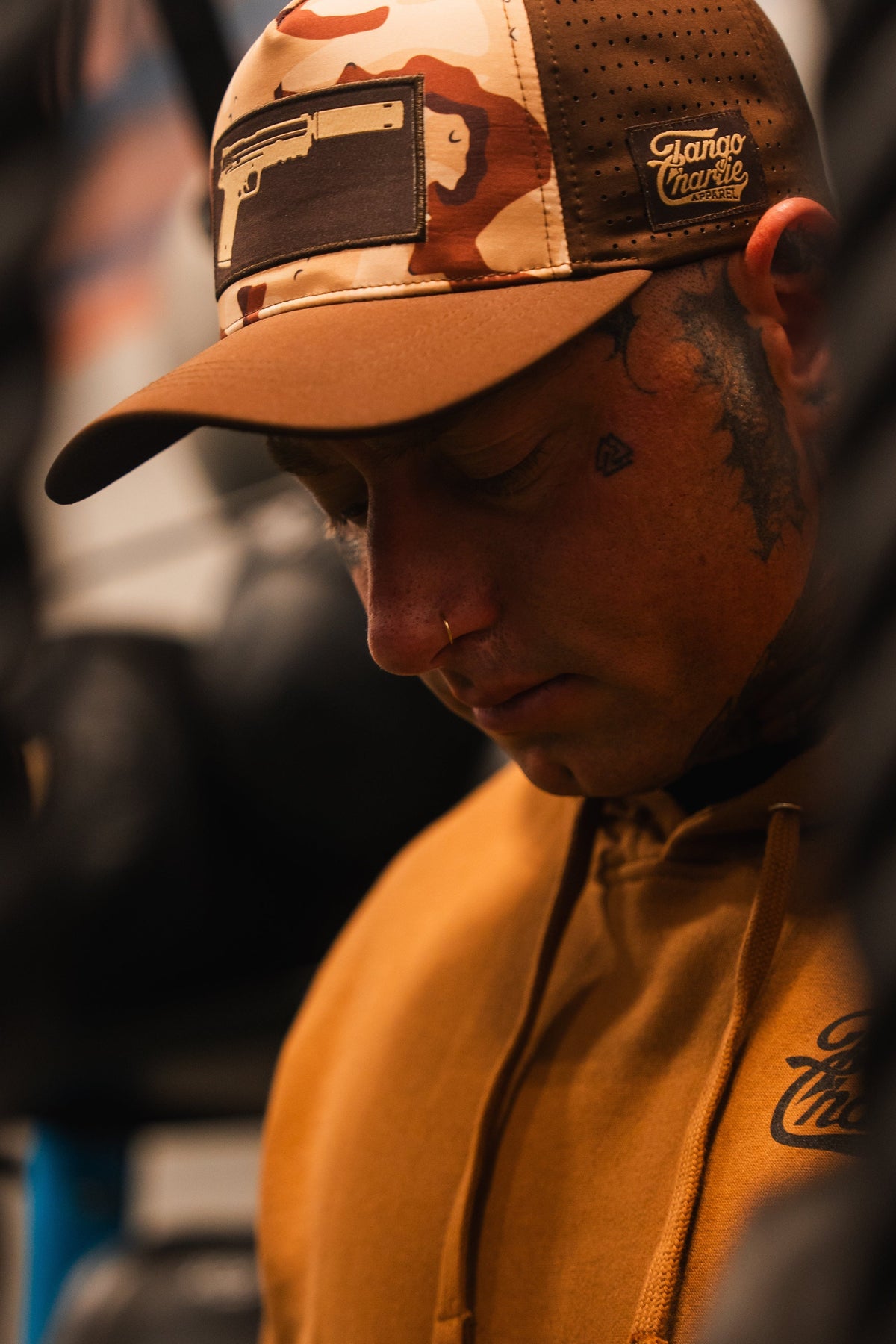 A person wears the Tango Charlie Apparel Choco Chip Silencer Hydro Snapback Hat, showcasing a gun graphic. The breathable, water-resistant snapback adds a modern edge, paired with facial tattoos and a nose ring against a blurred background.