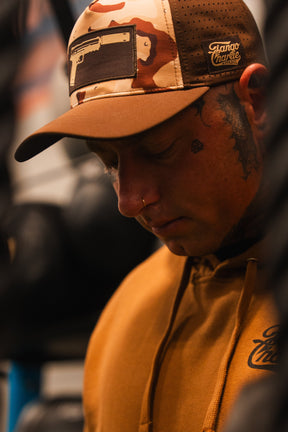A person wears the Tango Charlie Apparel Choco Chip Silencer Hydro Snapback Hat, showcasing a gun graphic. The breathable, water-resistant snapback adds a modern edge, paired with facial tattoos and a nose ring against a blurred background.