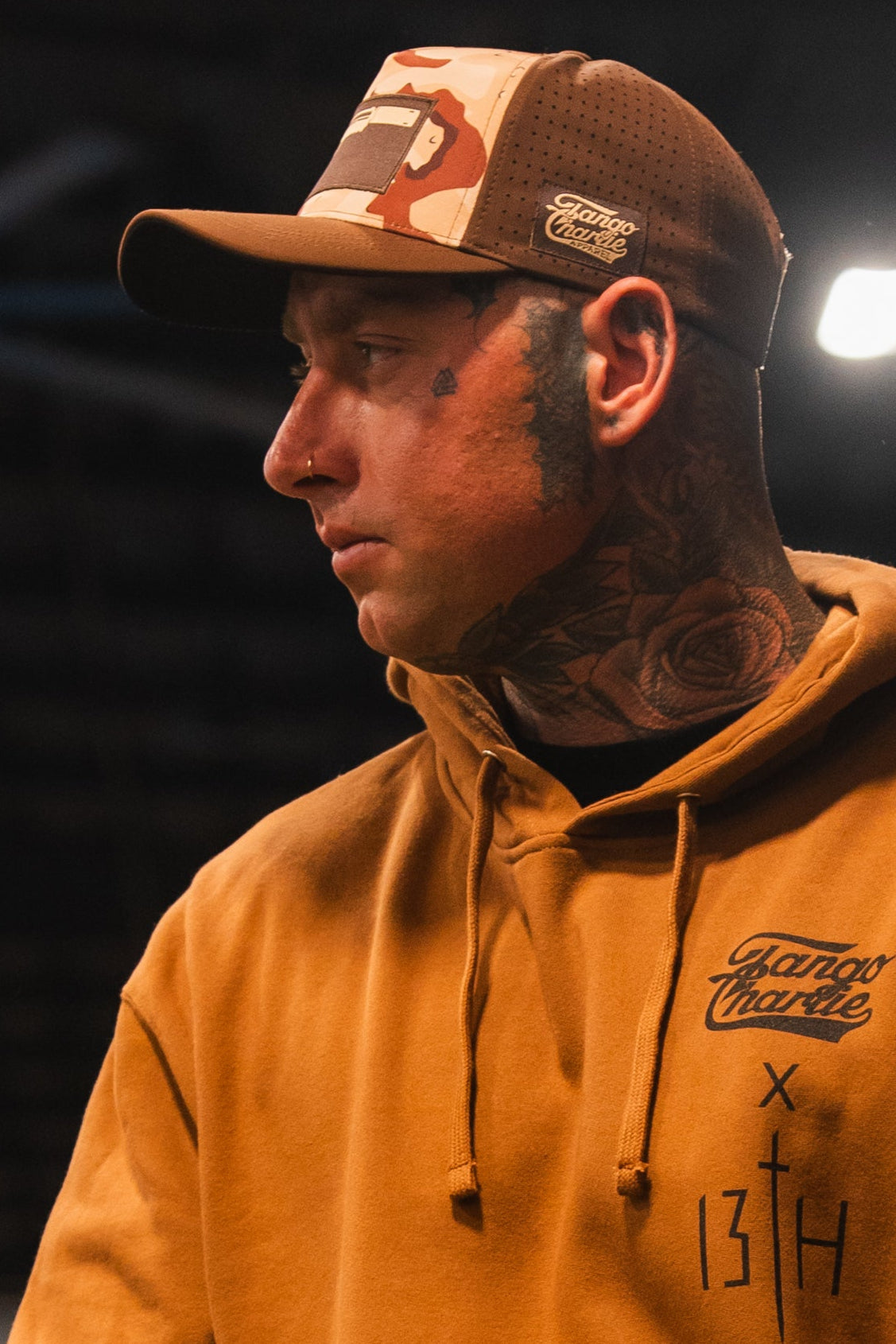 A man in a Tango Charlie Apparel Choco Chip Silencer Hydro Snapback Hat and tan hoodie, with face and neck tattoos, looks to the side. The background is dark and blurred with a bright light behind him.