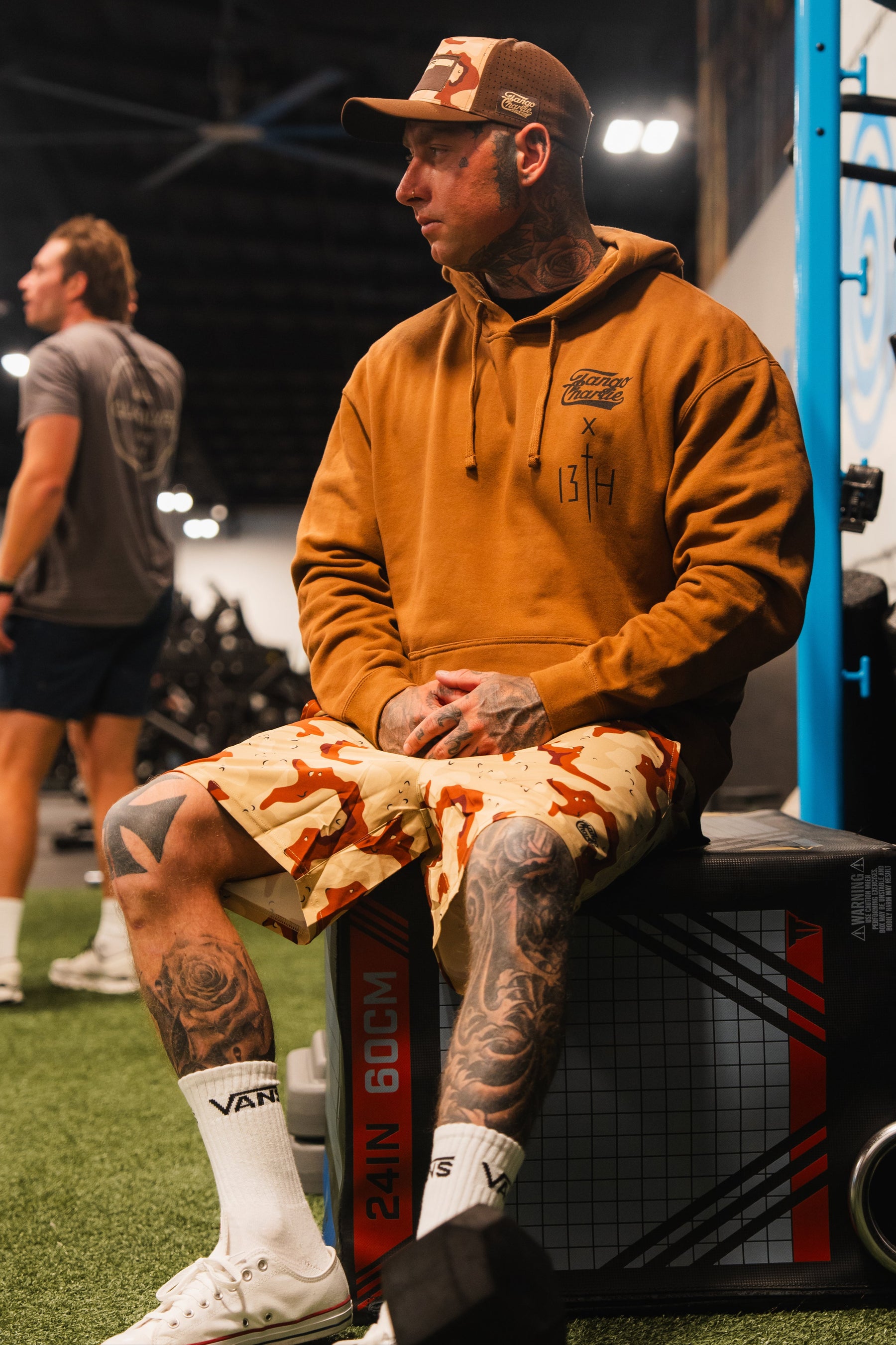 A tattooed man in a brown hoodie and Tango Charlie Apparel Men's Performance Training Shorts | 5.5"- Choco Chip sits on a gym plyo box, looking to the side with gym equipment and another person visible in the background.