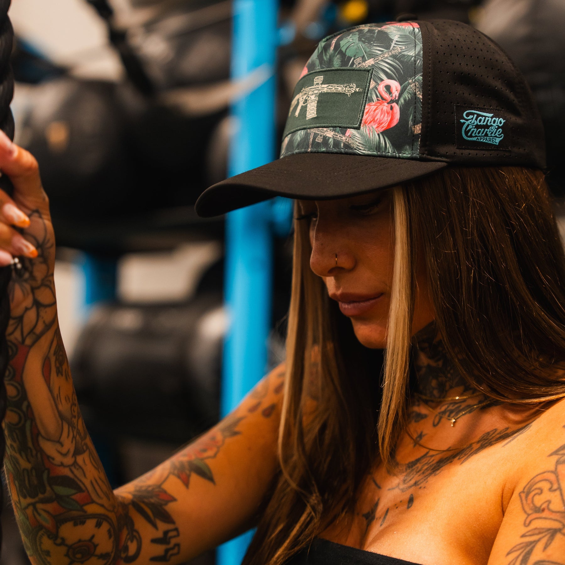 A person with long brown hair and tattooed arms wears the Loaded Flamingo Hydro Snapback Hat by Tango Charlie Apparel, featuring a black and green tropical print with a gun design, standing indoors near gym equipment.