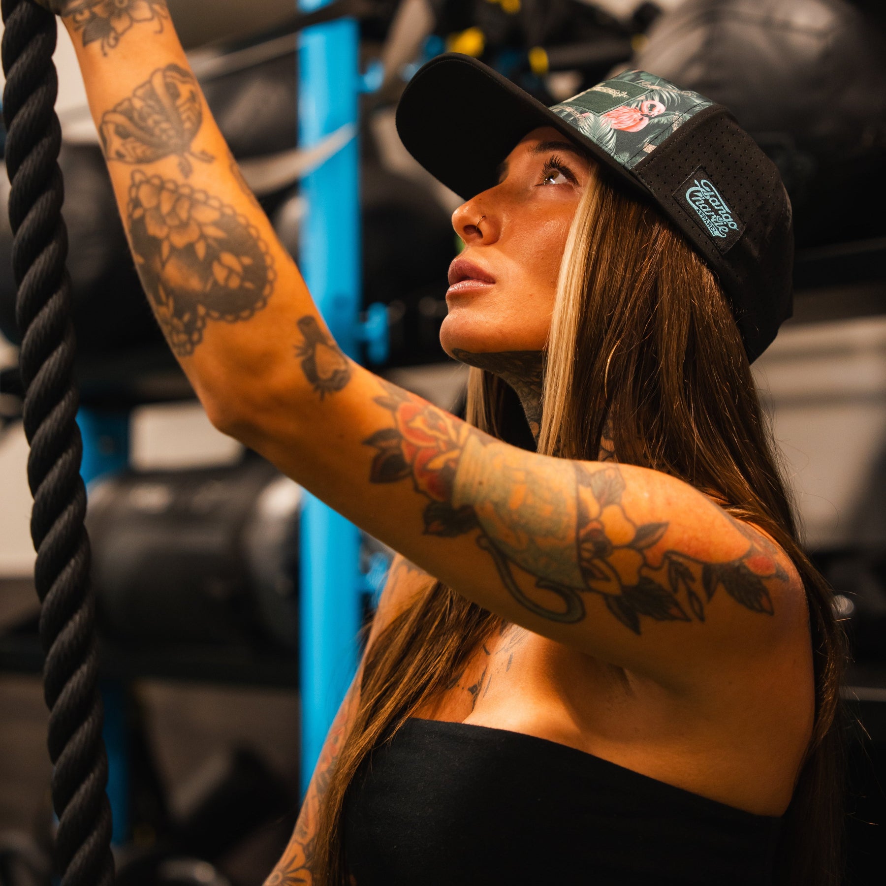 A woman with long hair and colorful arm tattoos wears the Loaded Flamingo Hydro Snapback Hat by Tango Charlie Apparel and a strapless top, looking upward and gripping a thick rope in a gym setting.