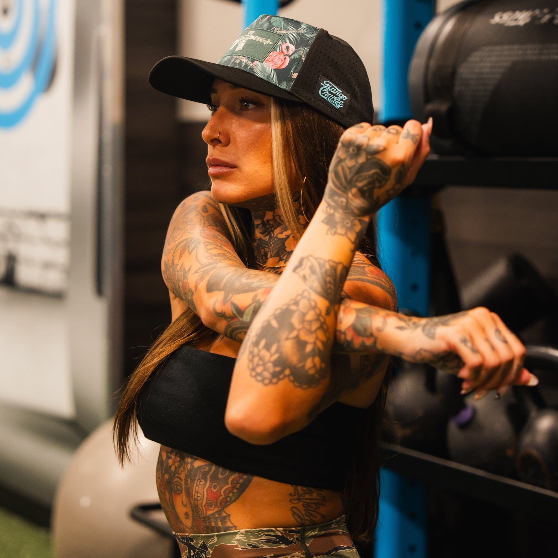 A tattooed woman wearing the Loaded Flamingo Hydro Snapback Hat by Tango Charlie Apparel and a tube top stretches her arm across her chest in a gym, with workout equipment visible in the background.