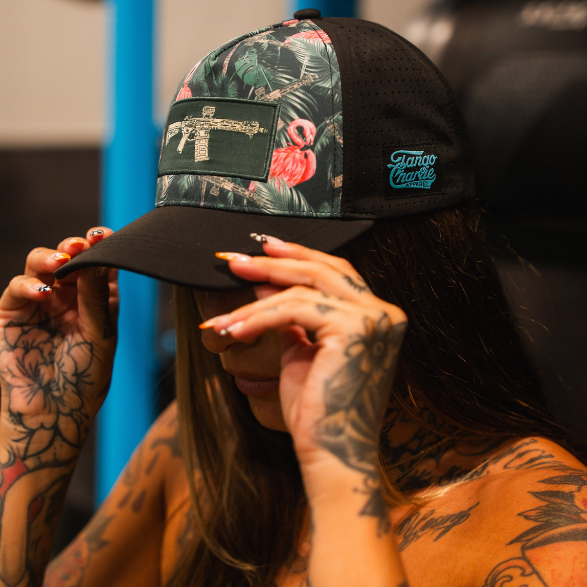 A tattooed woman adjusts a Loaded Flamingo Hydro Snapback Hat by Tango Charlie Apparel, featuring a floral flamingo design, submachine gun patch, and blue Tango Charlie logo. Her face is partially hidden.