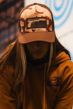 A person in a brown hoodie wears the Tango Charlie Apparel Choco Chip Silencer Hydro Snapback Hat featuring a black handgun graphic; their face is mostly shadowed and not fully visible.