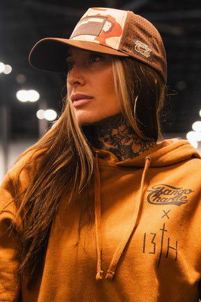 A woman wears a brown hoodie with "13TH" and "Bravo Charlie" prints, large hoop earrings, and the Tango Charlie Apparel Choco Chip Silencer Hydro Snapback Hat. She stands indoors with blurred lights, looking to the side, neck tattoos visible.