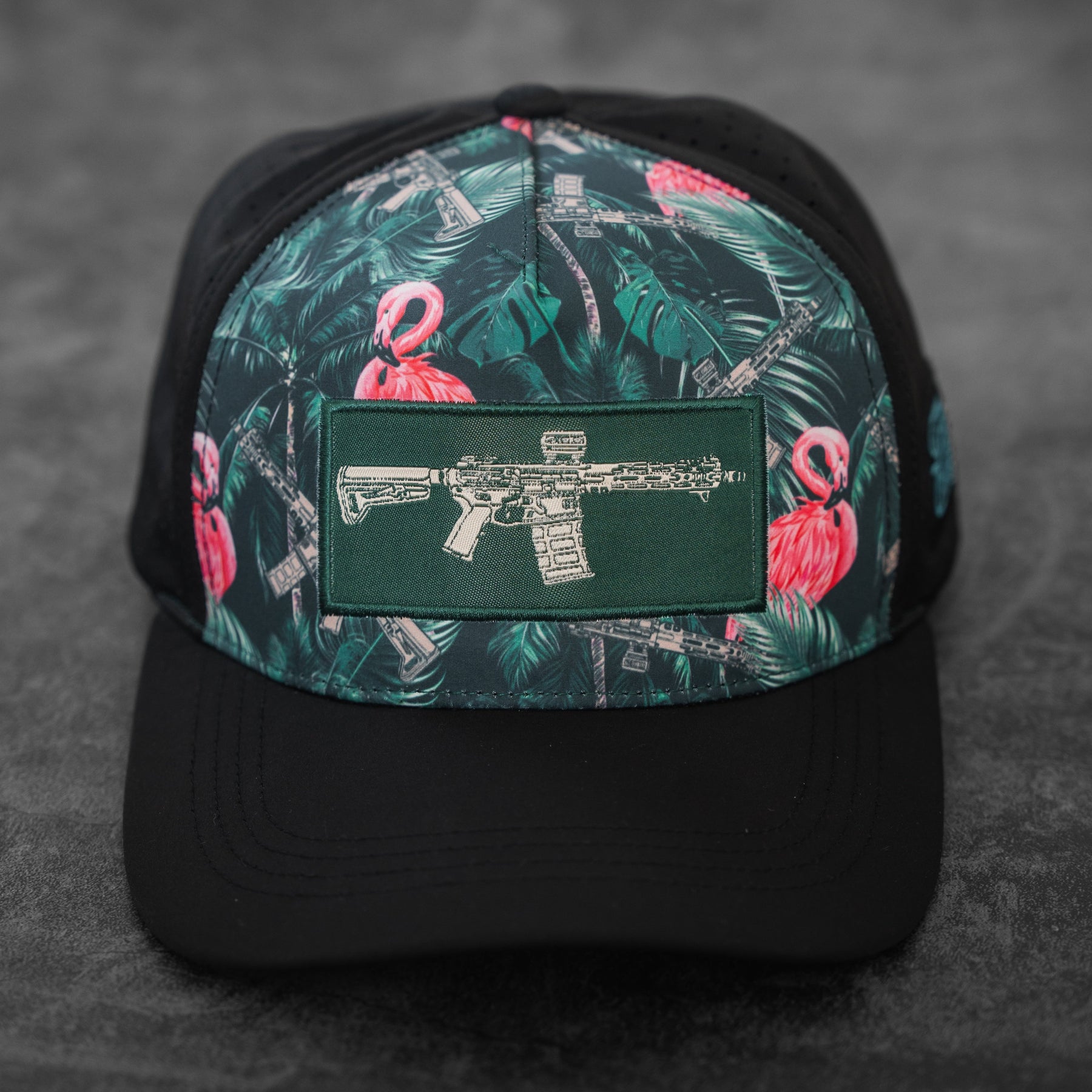 The Loaded Flamingo Hydro Snapback Hat by Tango Charlie Apparel features a black base with green and pink tropical flamingo and rifle patterns, plus a green patch displaying a rifle illustration. The cap is shown resting on a gray surface.