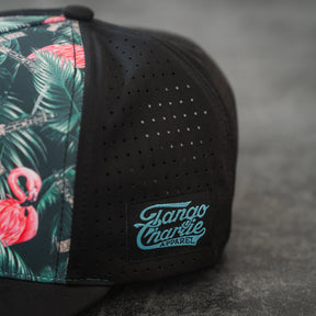 The Loaded Flamingo Hydro Snapback Hat by Tango Charlie Apparel features a black design with a vibrant flamingo and green leaf print on one panel, perforated ventilation holes, and a front Tango Charlie Apparel patch for style and comfort.