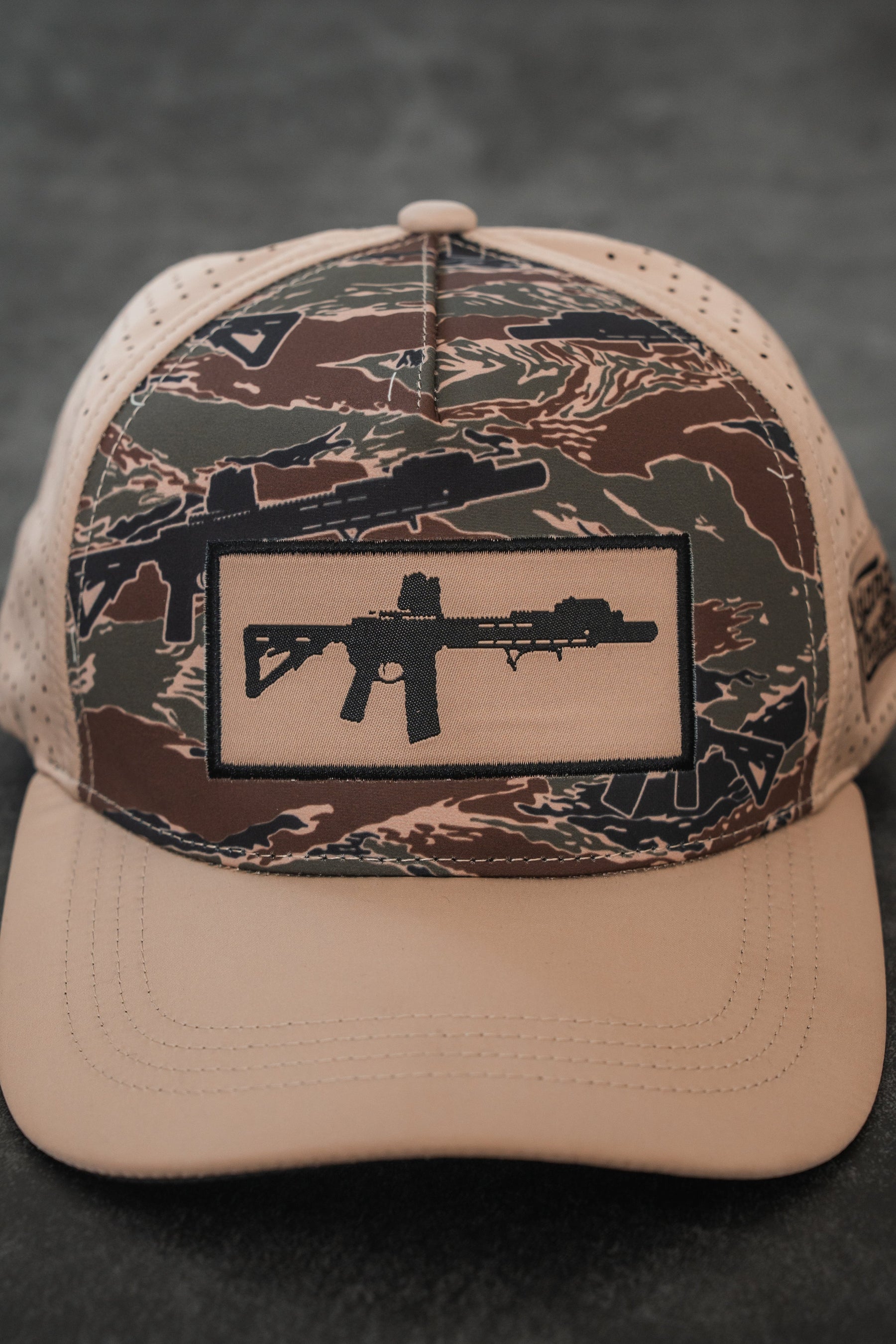 The Snipes & Stripes Hydro Snapback Hat by Tango Charlie Apparel features a camouflage pattern, tan brim, and a black patch with a modern rifle silhouette on the front.