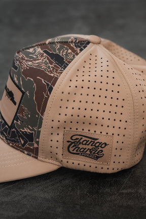 The Snipes & Stripes Hydro snapback hat by Tango Charlie Apparel features beige and camouflage colors, perforated side panels, and a branded patch on the side, displayed on a dark textured surface.