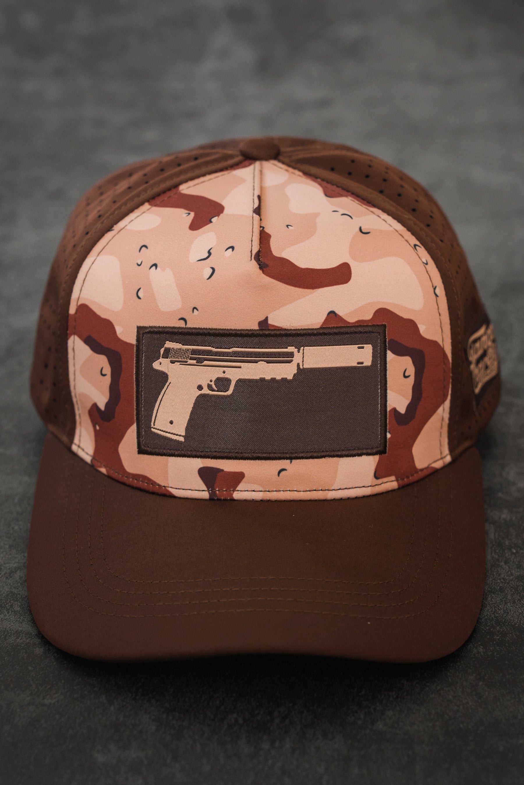 The Choco Chip Silencer Hydro Snapback Hat by Tango Charlie Apparel features a pistol with a silencer patch, brown brim, and perforated side panels for subtle style and functional detail.