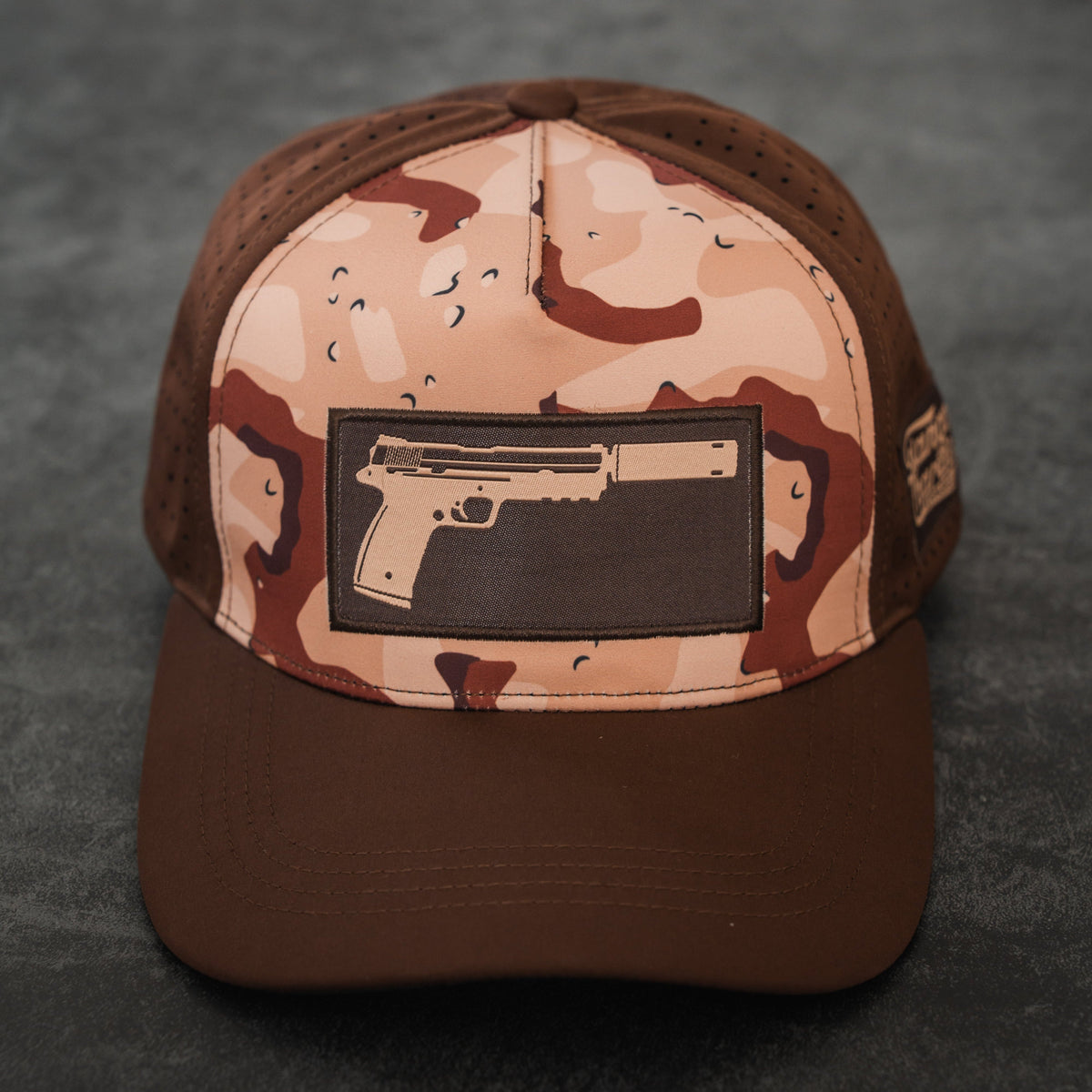 The Choco Chip Silencer Hydro Snapback Hat by Tango Charlie Apparel features brown and tan colors with a rectangular patch showing a handgun outline, displayed on a dark textured surface.