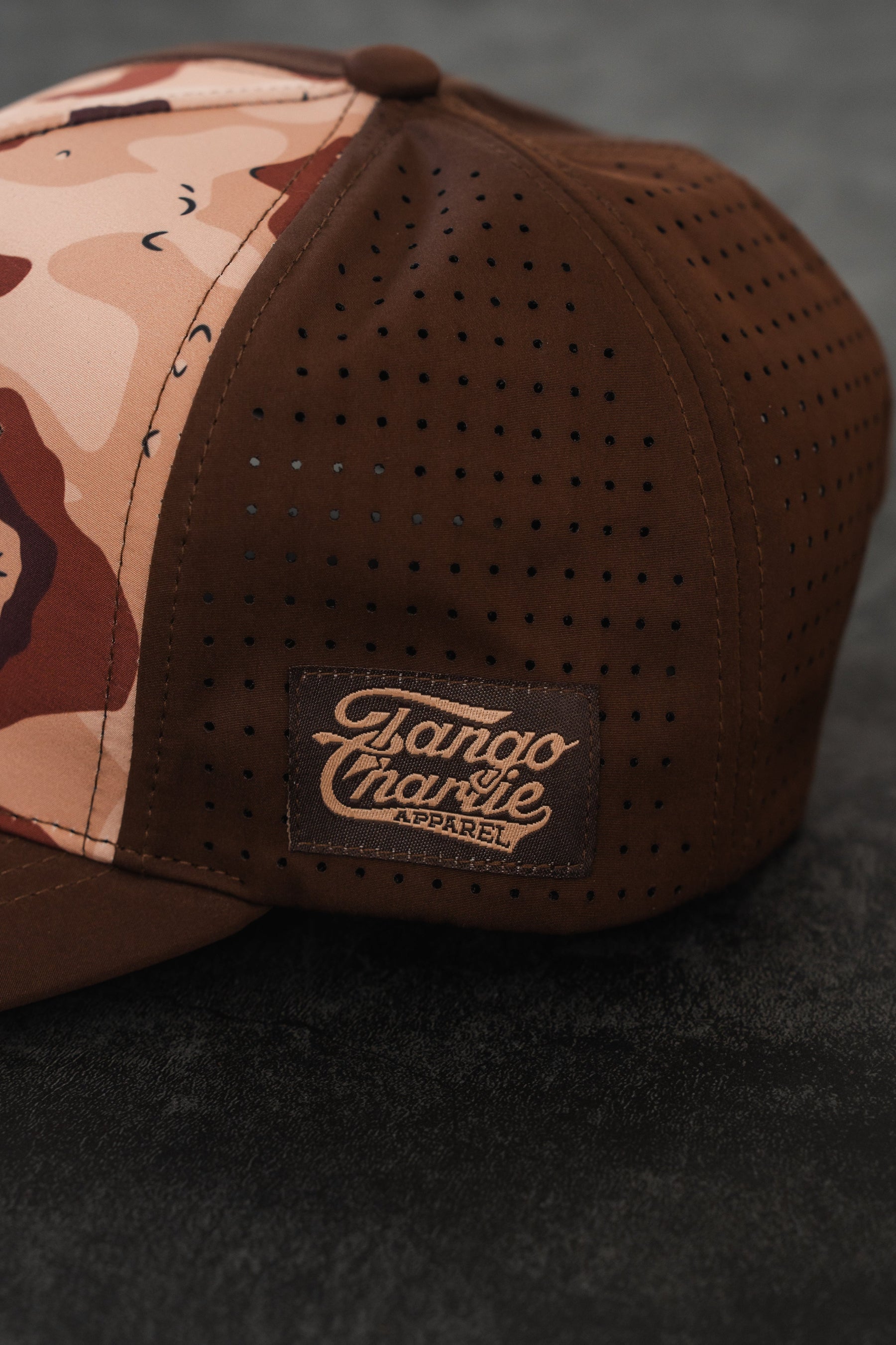 The Choco Chip Silencer Hydro Snapback Hat by Tango Charlie Apparel, featuring perforated side panels and a stitched brand patch, sits on a dark textured surface.