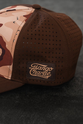 The Choco Chip Silencer Hydro Snapback Hat by Tango Charlie Apparel, featuring perforated side panels and a stitched brand patch, sits on a dark textured surface.