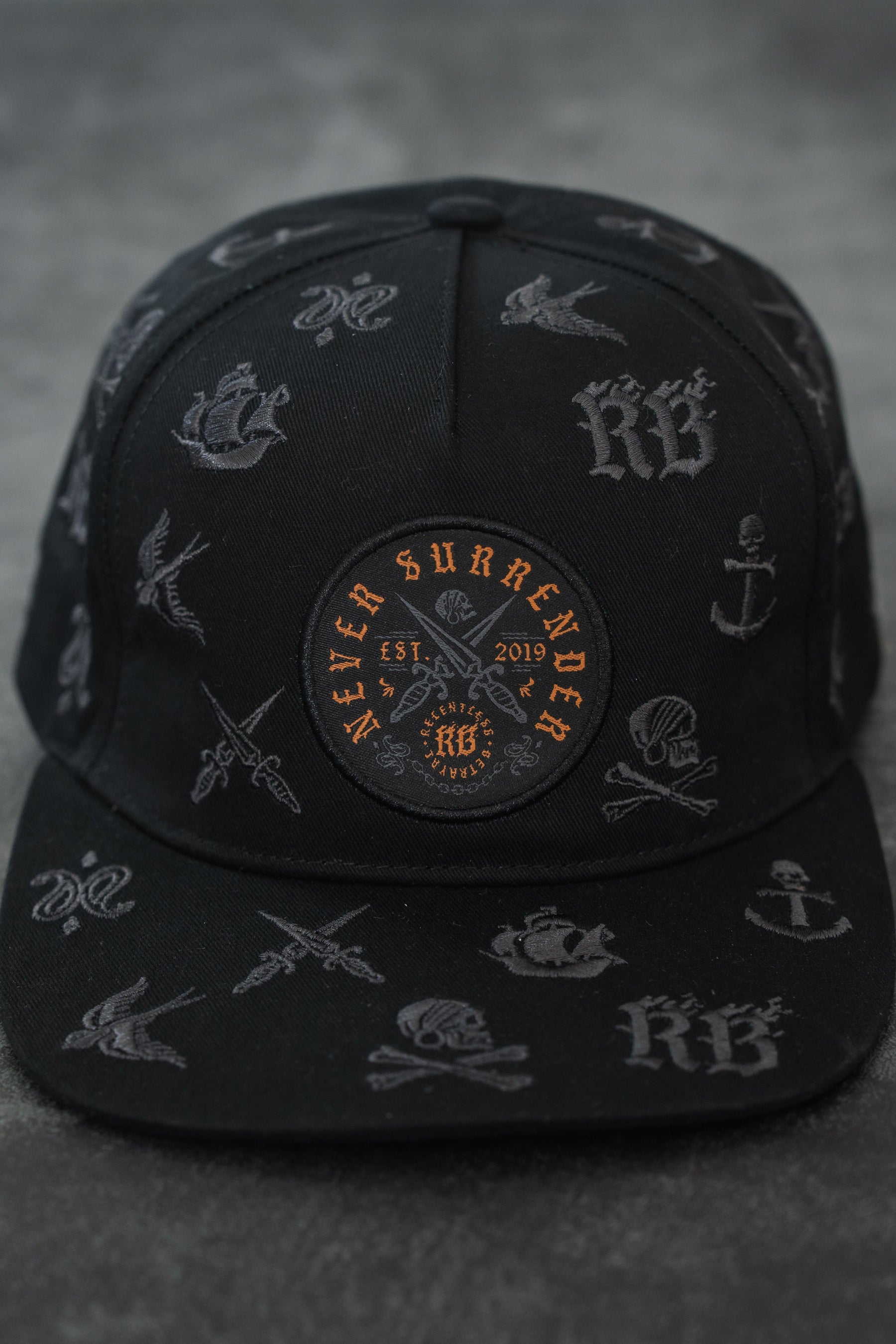 The Never Surrender Limited Edition Snapback by Relentless Betrayal is a black cap with grey nautical and tattoo-style designs, plus an orange and black patch on the front reading "Never Surrender Est. 2019.